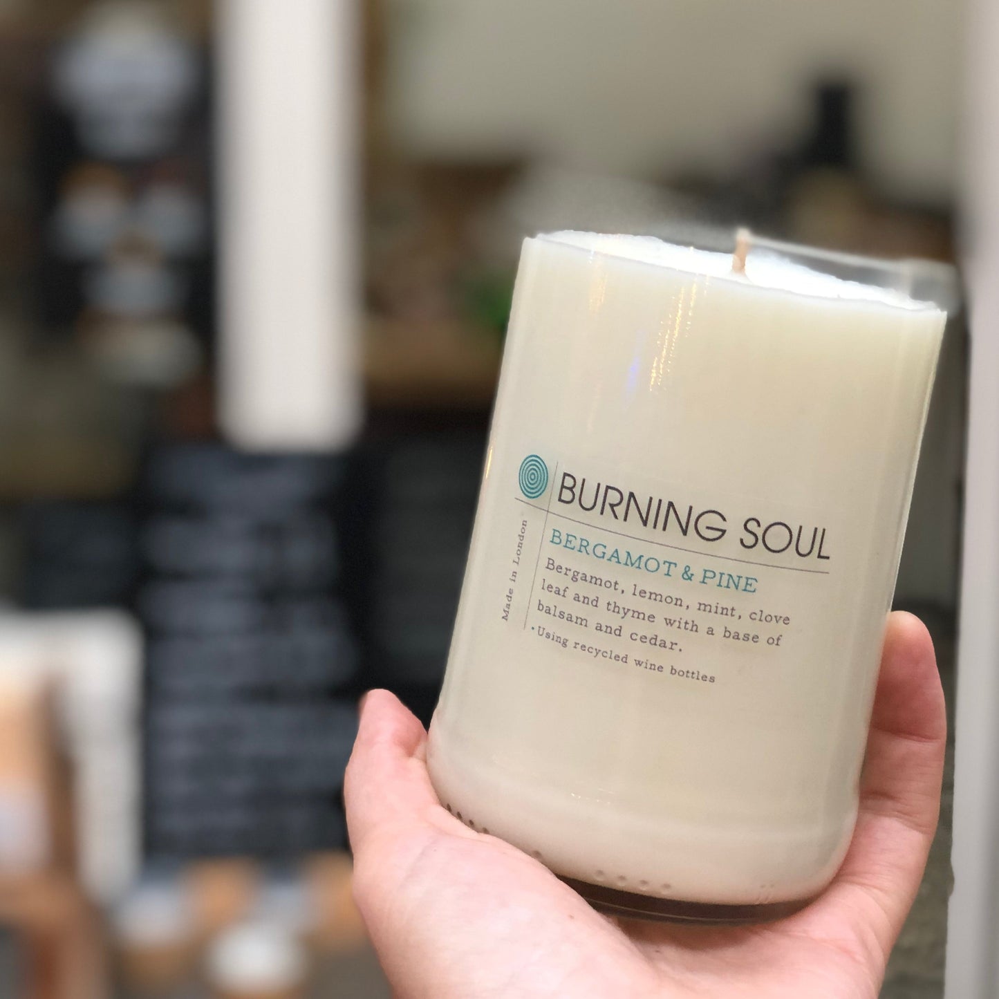 hand holding up a white burning soul candle in an upcycled wine bottle, background is blurred weigh and pay shop, bergamot pine scent