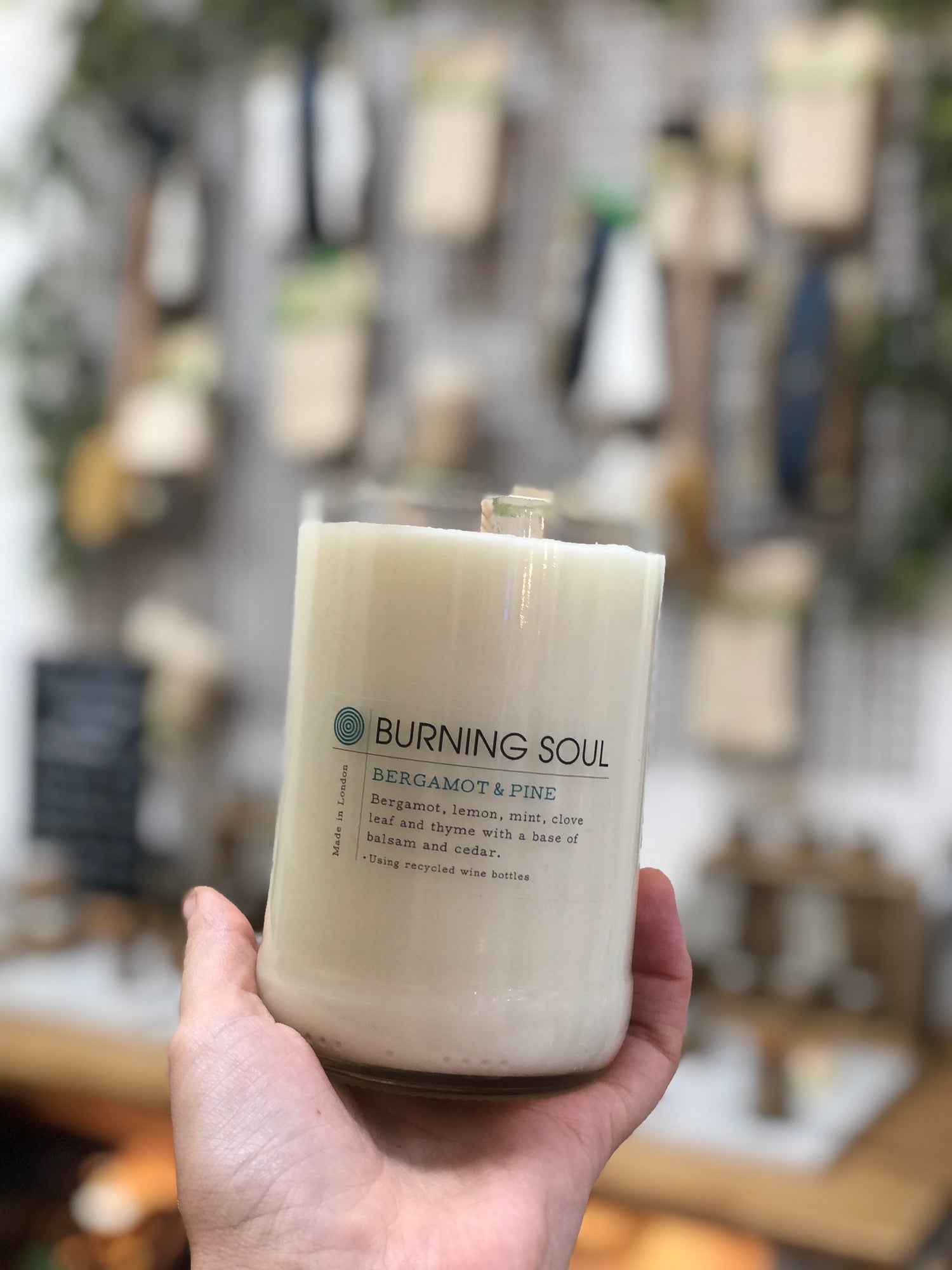 hand holding a wine bottle candle in bergamot and pine by burning soul, with blurred weigh and pay shop in background